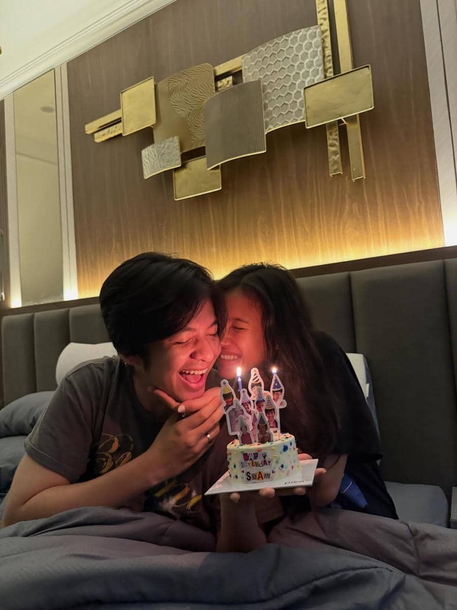 Sepasang anak muda tertawa bahagia di atas tempat tidur sambil memegang kue ulang tahun kecil dengan dua lilin menyala.