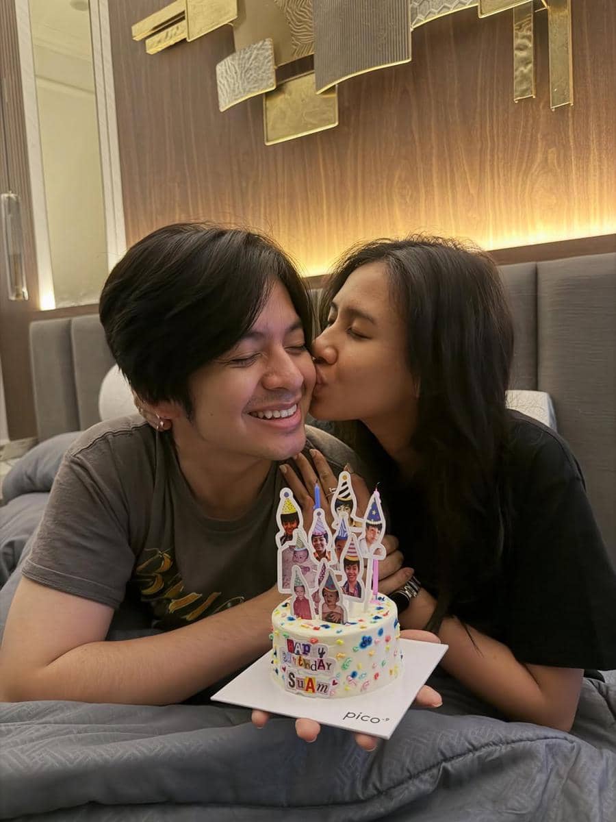 Seorang perempuan mencium pipi seorang pria yang tersenyum sambil memegang kue ulang tahun kecil dengan hiasan foto di atasnya.