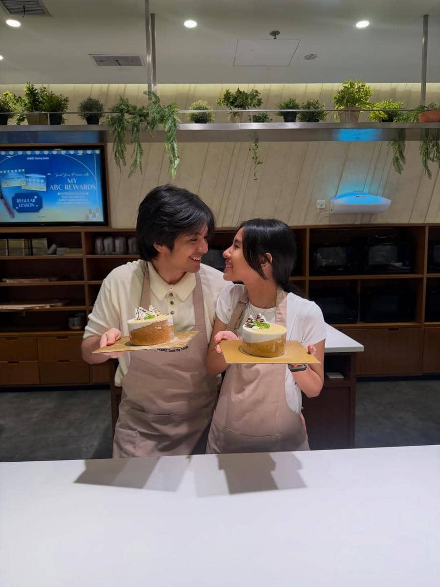 Sepasang pria dan wanita mengenakan celemek sambil tersenyum dan memegang kue di dapur modern dengan tanaman hias di rak atas.