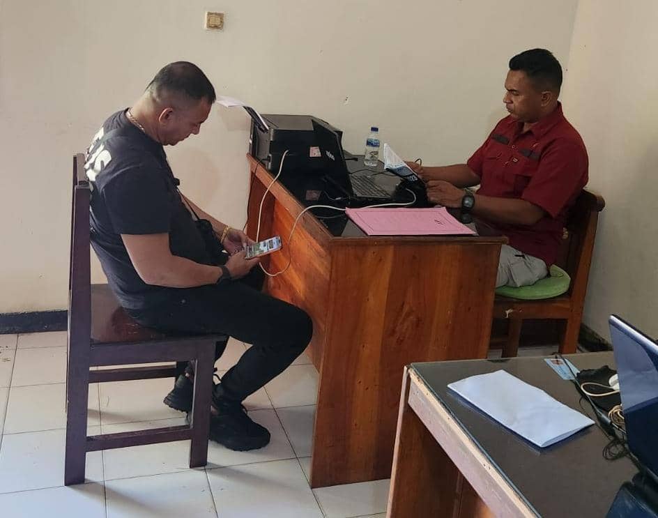 Seorang pria duduk di kursi sambil memegang ponsel di hadapan petugas yang bekerja di meja kantor dengan komputer dan berkas.