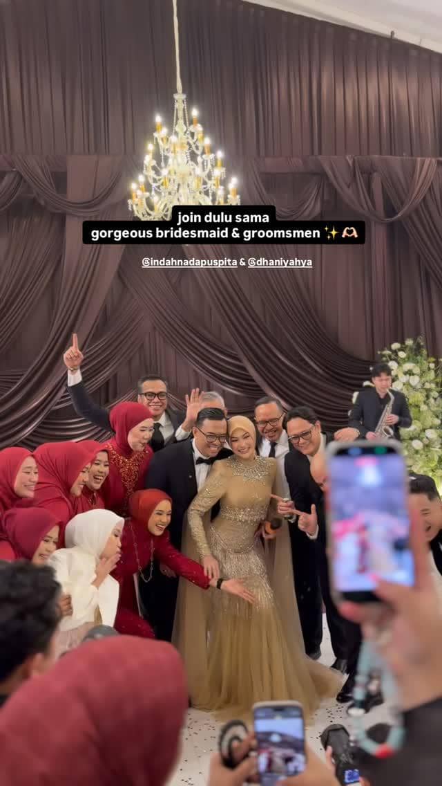 Pengantin berpose bersama bridesmaid dan groomsmen di acara pernikahan dengan latar tirai cokelat dan lampu gantung mewah.