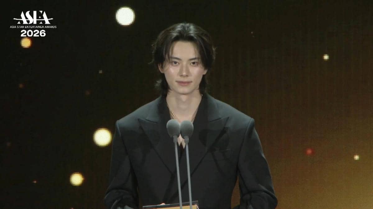 Seorang pembawa acara mengenakan setelan hitam berdiri di depan mikrofon ganda di panggung Asia Star Entertainer Awards 2026.