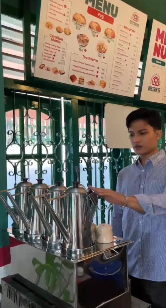 Seorang pria mengenakan kemeja biru sedang menyiapkan minuman di area penyajian kopi dengan deretan teko logam di kafe Distrik15 Kota Metro.
