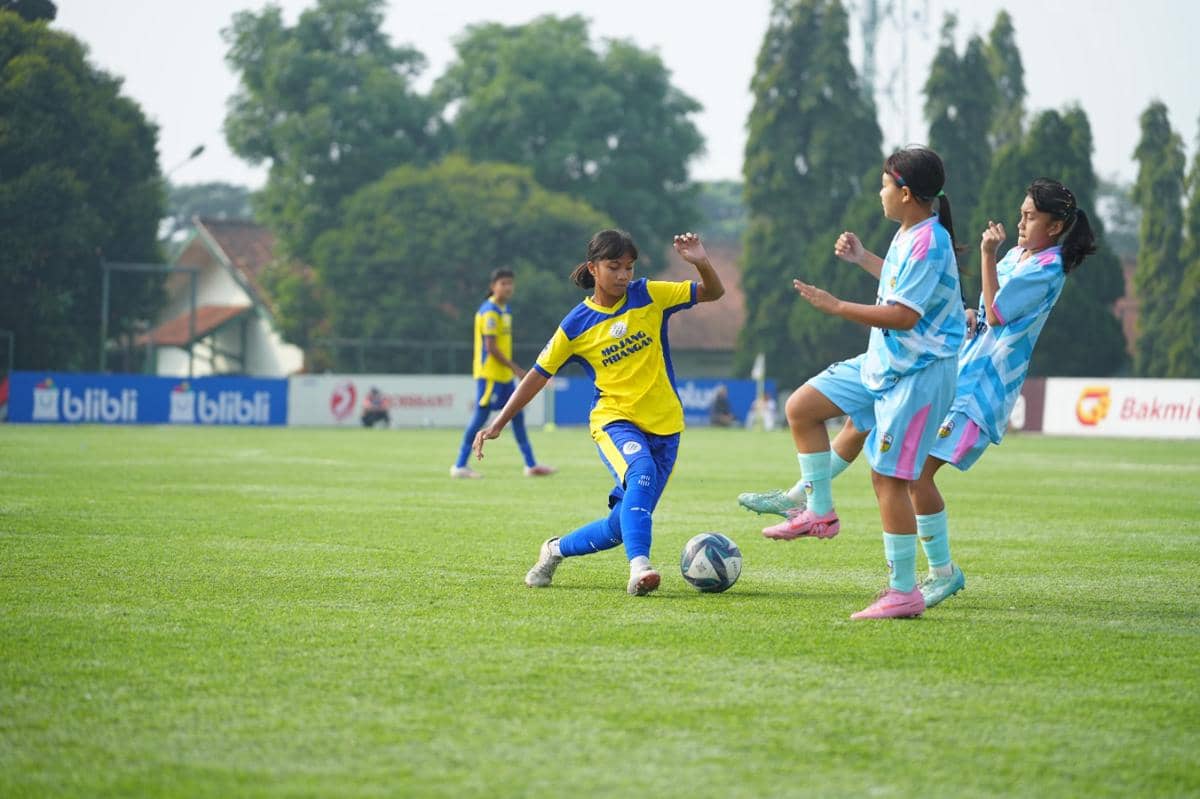 Pemain muda mengenakan seragam kuning biru menggiring bola menghadapi dua lawan berseragam biru muda di lapangan sepak bola.