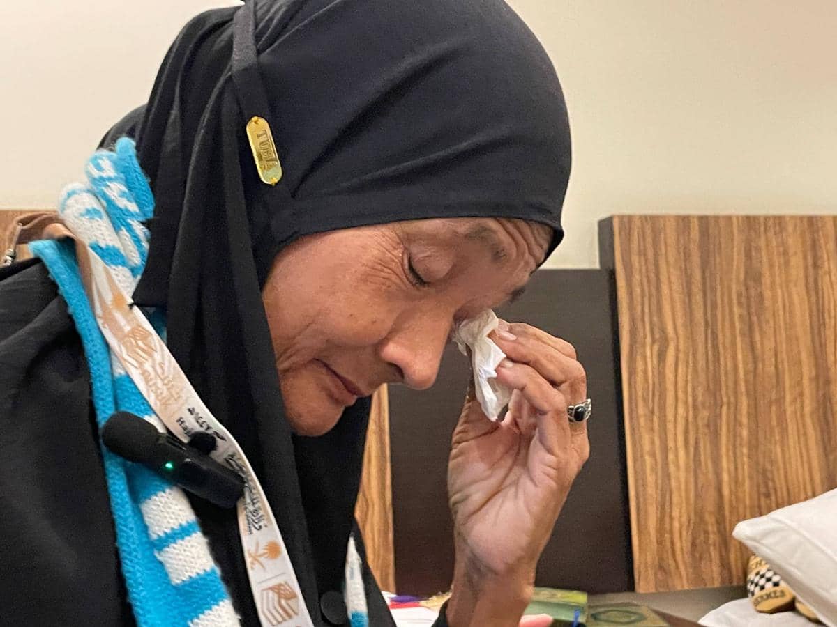 Seorang perempuan lanjut usia mengenakan hijab hitam dan syal biru putih sedang menyeka air mata dengan tisu di dalam ruangan.