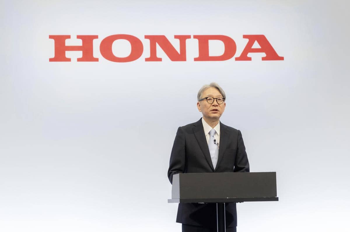Seorang eksekutif Honda berdiri di podium mengenakan setelan jas dan dasi, berbicara di depan logo besar Honda berwarna merah.