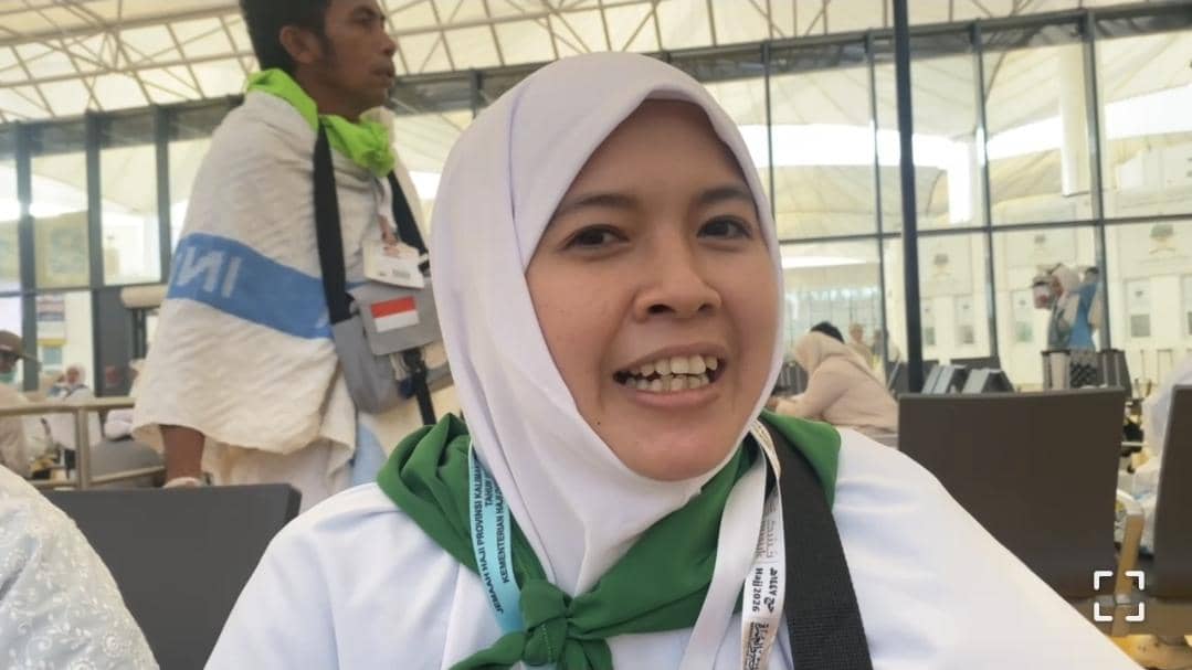 Seorang jemaah haji perempuan mengenakan hijab putih dan syal hijau tersenyum di area tunggu embarkasi dengan latar jamaah lain.