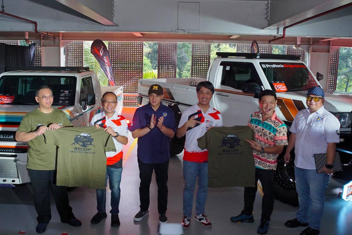 Beberapa pria berpose di depan mobil modifikasi sambil memegang kaus bertema otomotif di sebuah acara komunitas pecinta mobil.