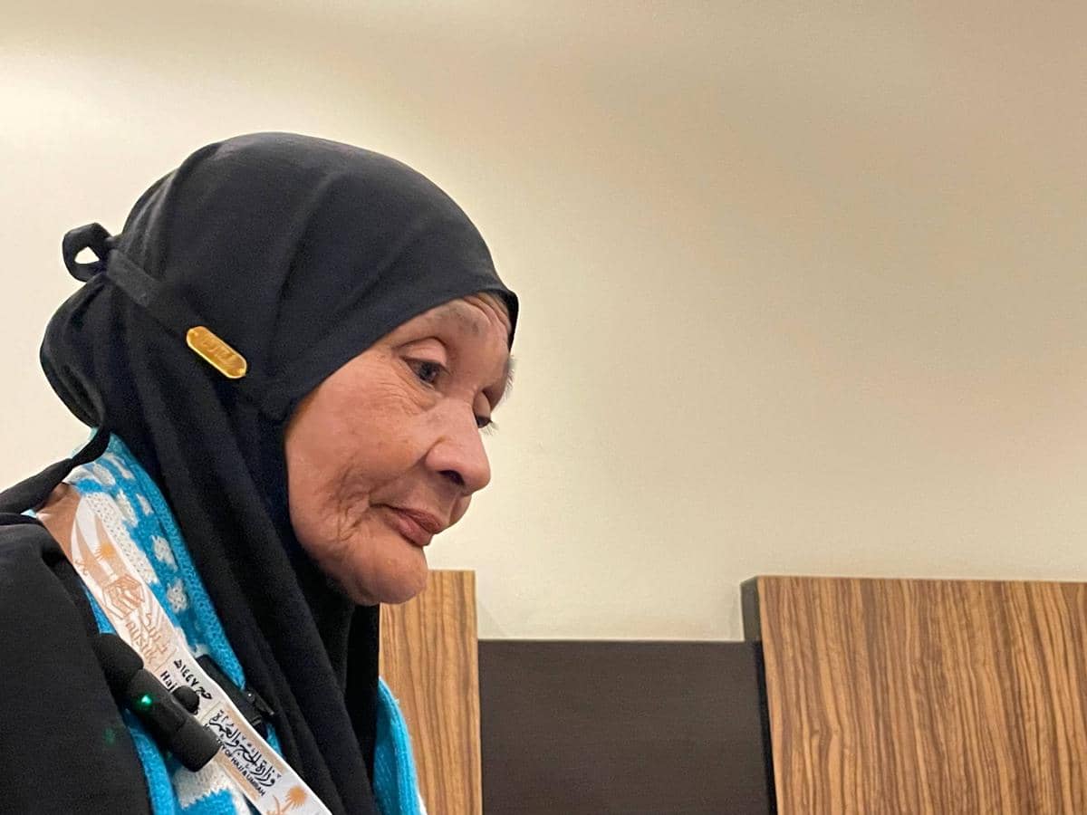 Seorang perempuan lanjut usia mengenakan hijab hitam dan syal biru putih dengan latar ruangan sederhana berwarna krem.