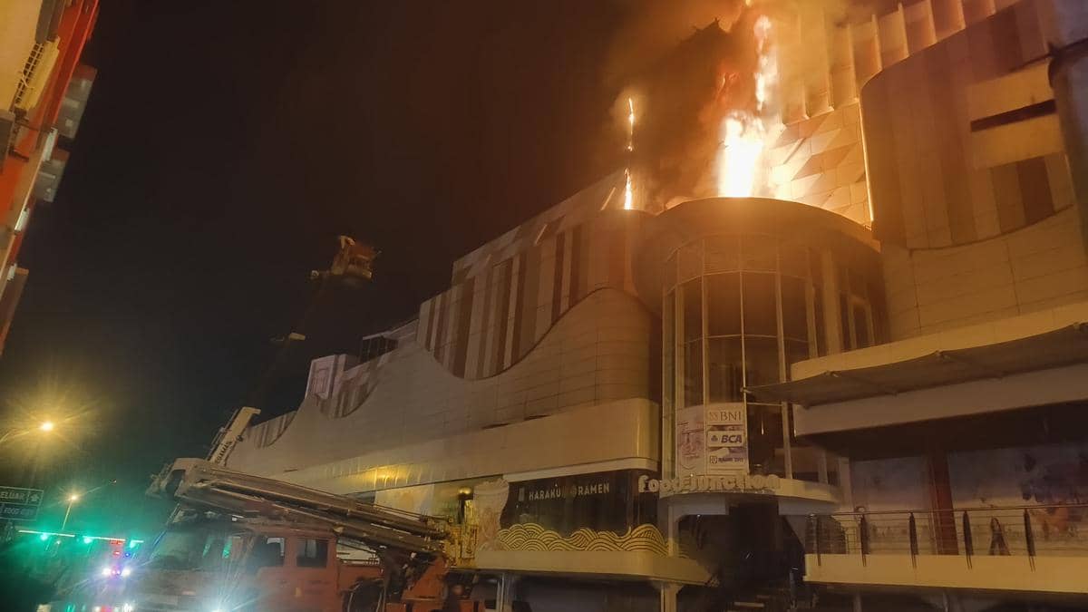 Api besar membakar bagian atas gedung Mega Mall Manado pada malam hari, dengan mobil pemadam kebakaran beroperasi di lokasi.