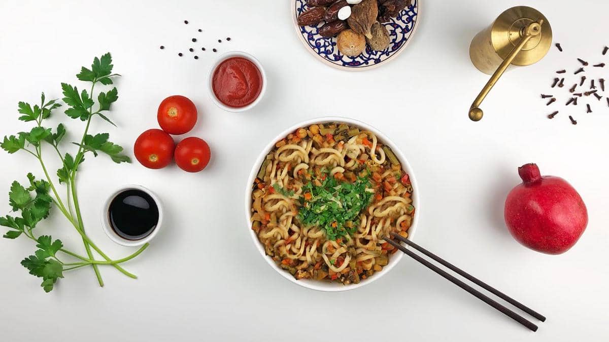 Semangkuk lagman dengan potongan sayuran berwarna-warni, dihiasi daun ketumbar, dikelilingi tomat, saus, dan bumbu di atas meja putih.