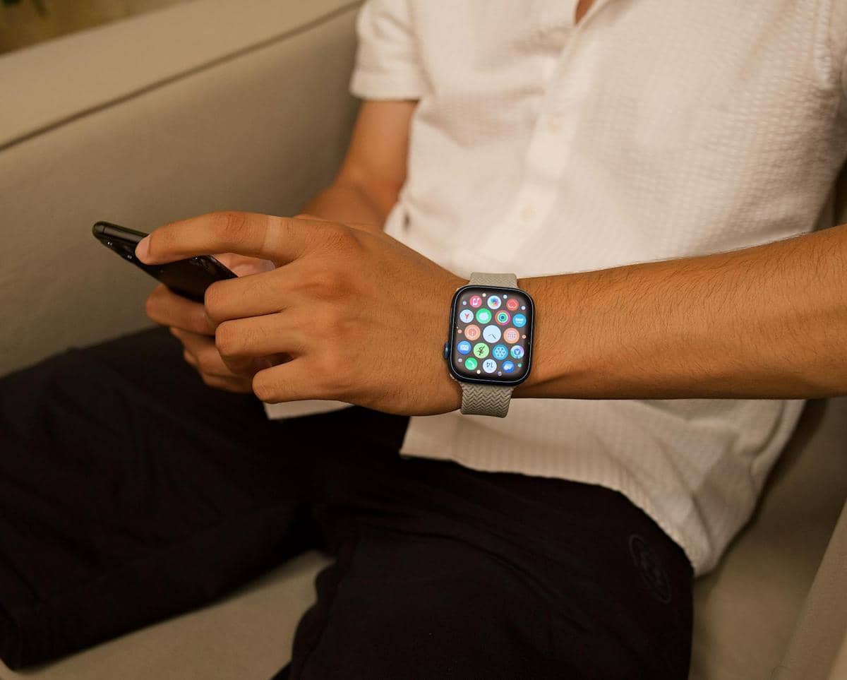 Seseorang mengenakan smartwatch dengan layar berwarna cerah di pergelangan tangan sambil memegang ponsel di sofa.