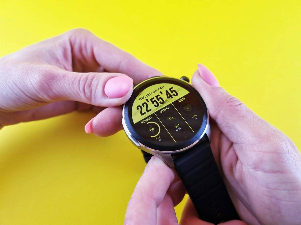 Seseorang memegang smartwatch dengan layar digital berwarna kuning yang menampilkan waktu dan data langkah di latar belakang kuning cerah.