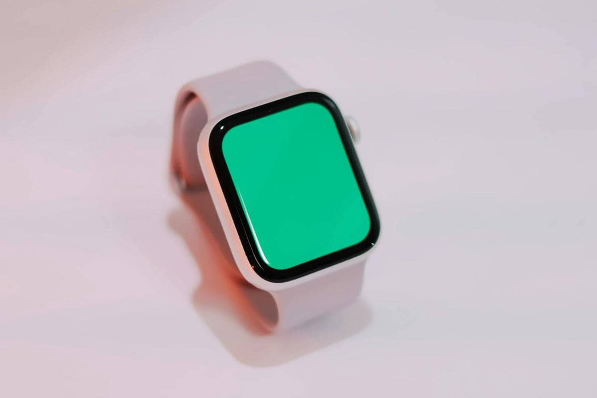 Smartwatch berwarna putih dengan layar hijau menyala diletakkan di permukaan datar dengan pencahayaan lembut.