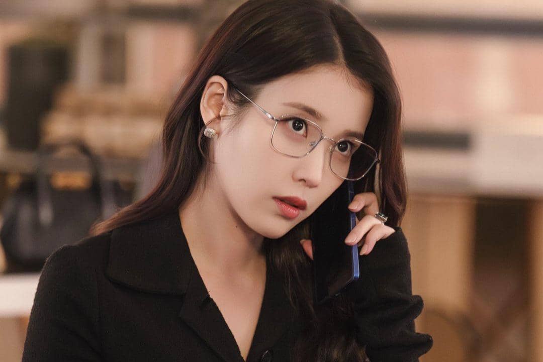 Seorang wanita berambut panjang mengenakan kacamata dan pakaian hitam sedang berbicara di telepon dalam adegan drama Perfect Crown.