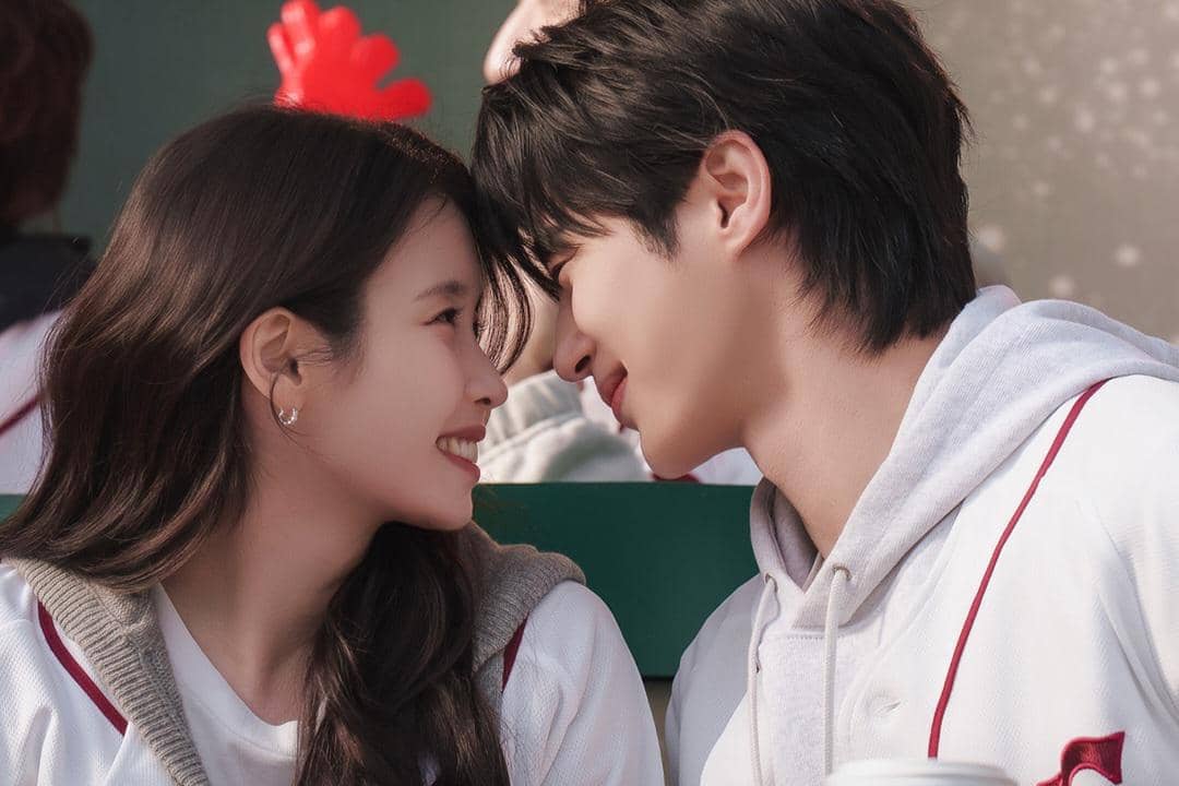Dua karakter drama Perfect Crown saling menatap dengan senyum hangat, mengenakan pakaian kasual bernuansa putih dan merah.