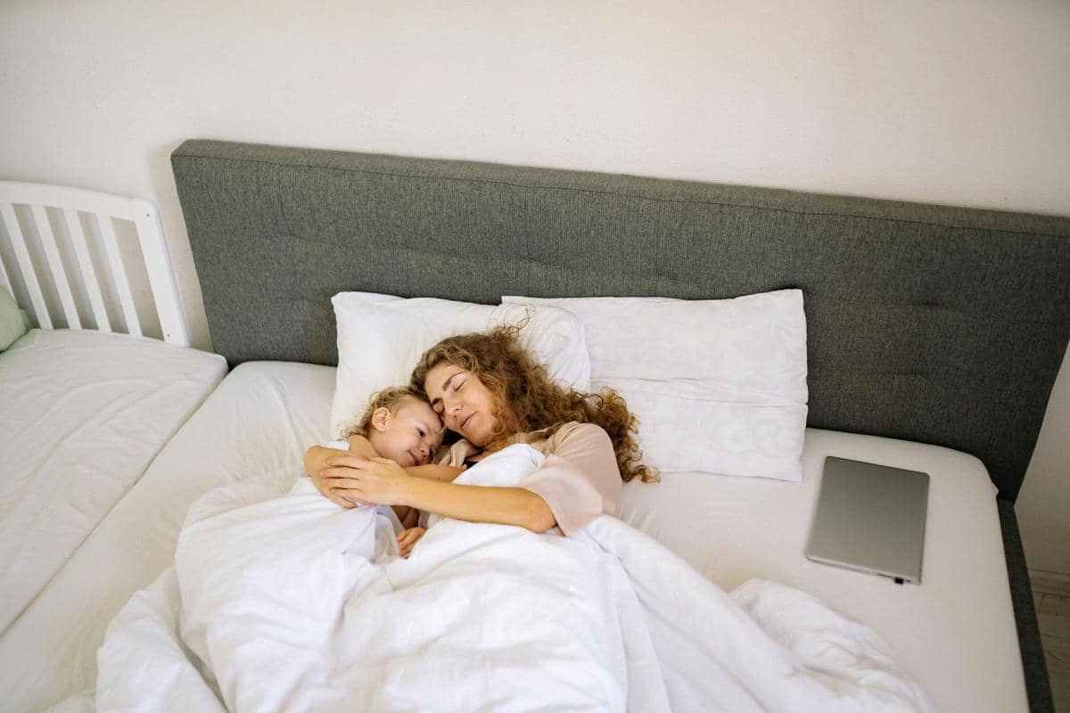Seorang ibu dan anak kecil tidur bersama di tempat tidur dengan selimut putih, tampak tenang dan hangat di kamar yang terang.