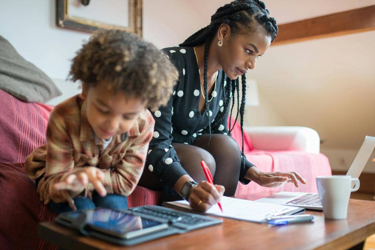 Seorang ibu bekerja di rumah menggunakan laptop sambil menulis catatan, sementara anaknya bermain dengan tablet di meja ruang tamu.