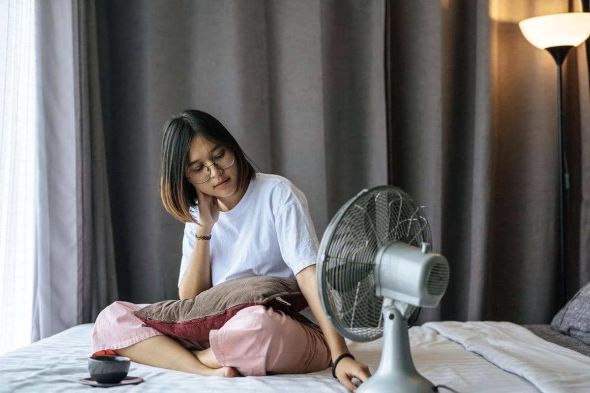 Seorang perempuan duduk di tempat tidur menyalakan kipas angin di kamar dengan tirai tertutup saat cuaca panas.