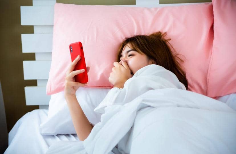 Seorang wanita berbaring di tempat tidur dengan bantal merah muda sambil melihat ponsel berwarna merah setelah bangun tidur.