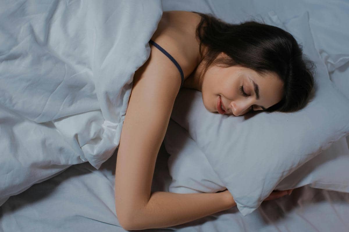 Seorang wanita tidur nyenyak di atas tempat tidur dengan selimut putih dan bantal, tampak tenang dan nyaman di kamar tidur.