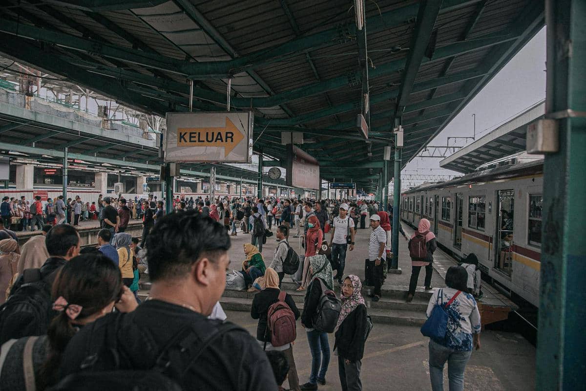 Suasana ramai di stasiun kereta dengan banyak penumpang menunggu dan berjalan di peron dekat kereta yang sedang berhenti.