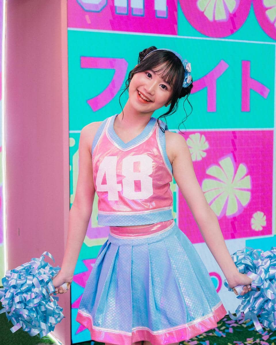 Seorang perempuan mengenakan kostum berwarna pink dan biru dengan angka 48 di bagian depan, memegang pom-pom di kedua tangan dan tersenyum di depan latar warna-warni.
