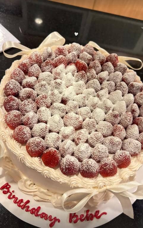 Kue ulang tahun bertopping stroberi segar dan taburan gula halus dengan tulisan ucapan di bagian bawah kue.