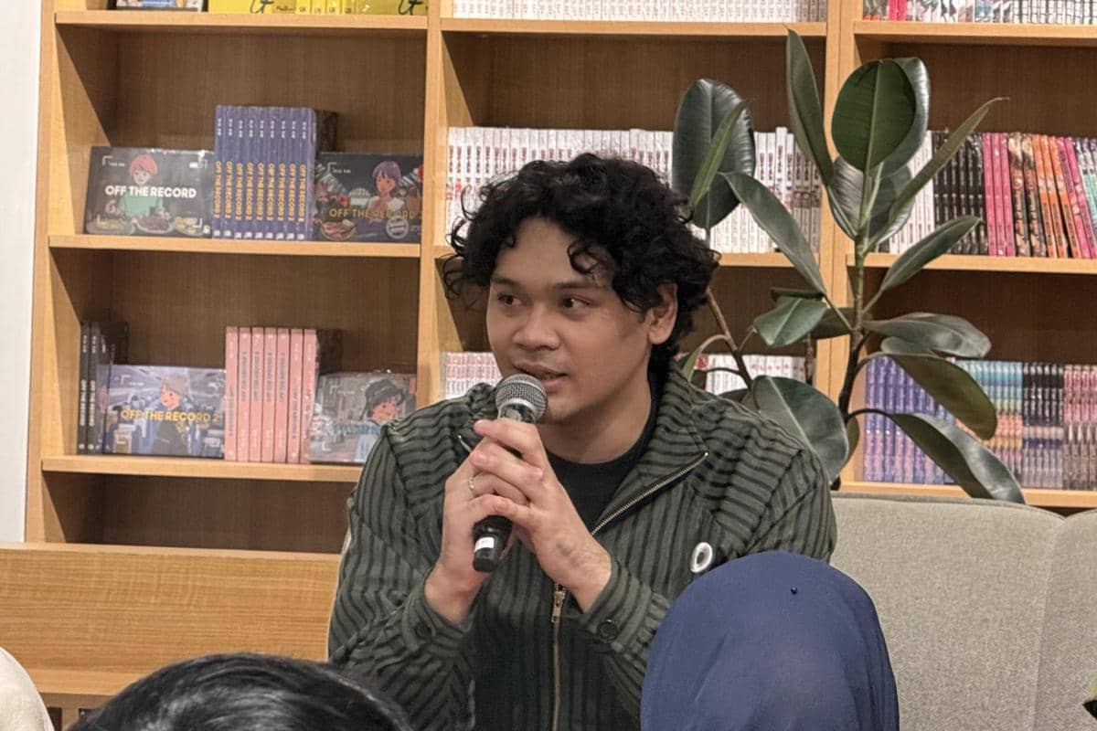 Seorang pria berbicara memegang mikrofon saat konferensi pers The Overtunes, dengan latar rak buku di sebuah ruangan.