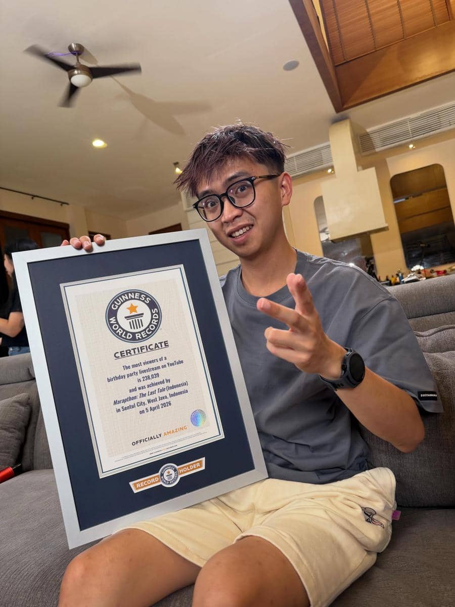 Seorang pria muda duduk di sofa sambil memegang sertifikat Guinness World Records berbingkai besar di ruang tamu modern.