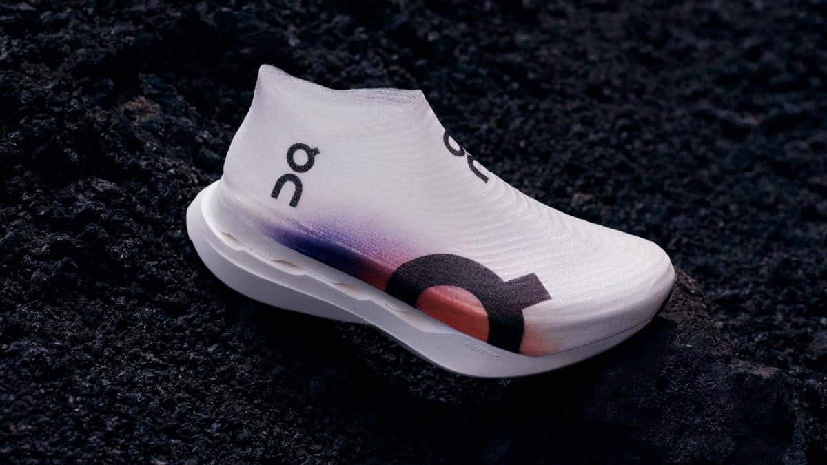 Sepatu lari On Cloudboom Strike LS berwarna putih dengan gradasi ungu dan oranye, diletakkan di permukaan hitam bertekstur kasar.