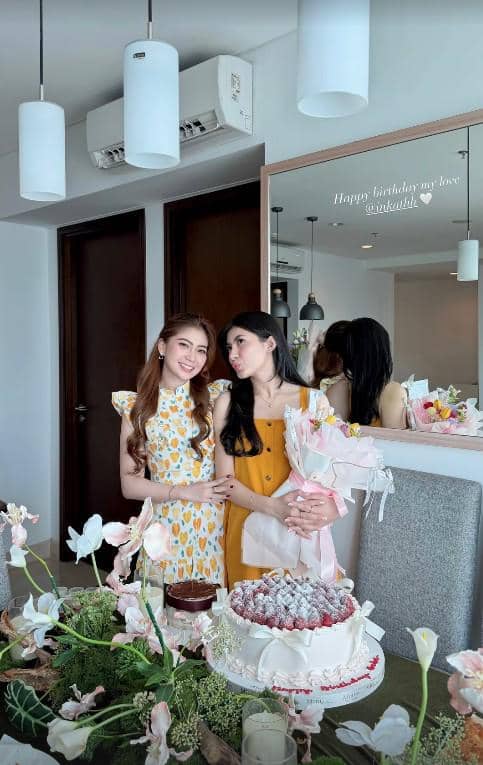 Dua wanita berpose di depan meja dengan kue ulang tahun dan bunga, salah satunya memegang buket bunga di ruangan bernuansa cerah.