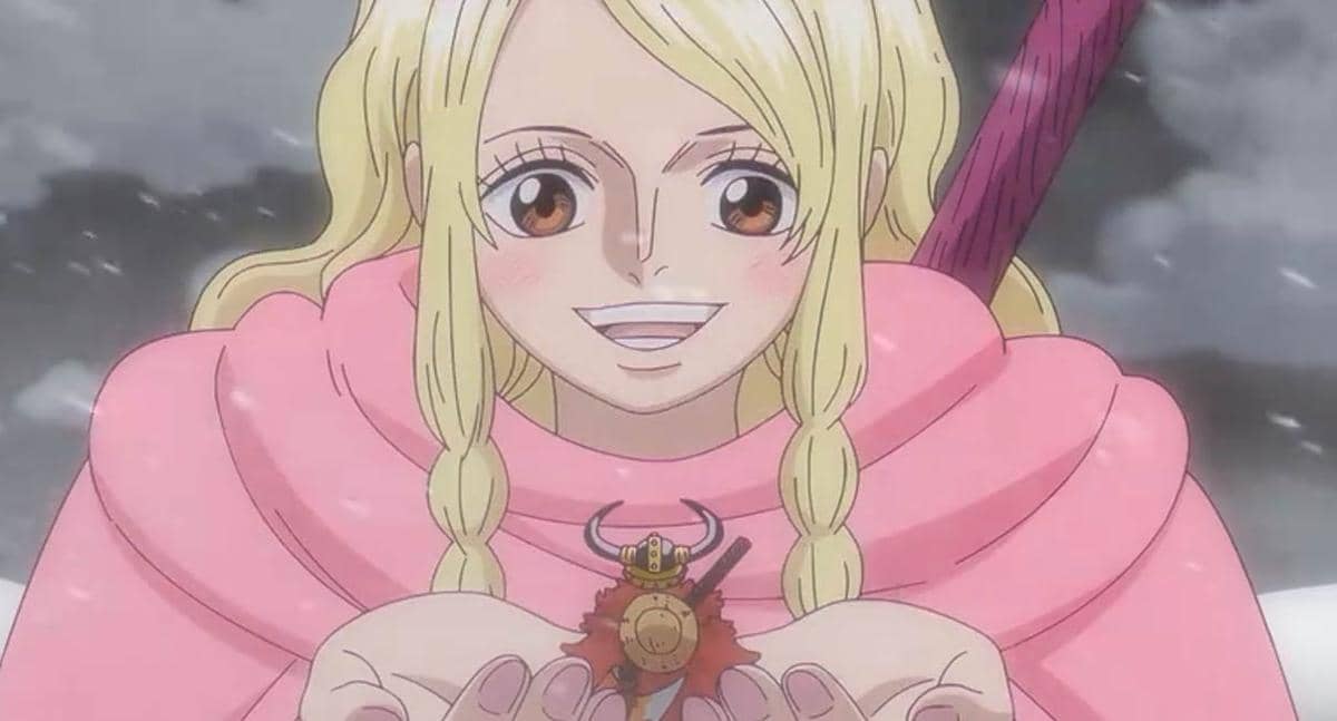 Gerd berambut pirang tersenyum sambil memegang miniatur Luffy di tangannya, mengenakan jubah merah muda di tengah salju.