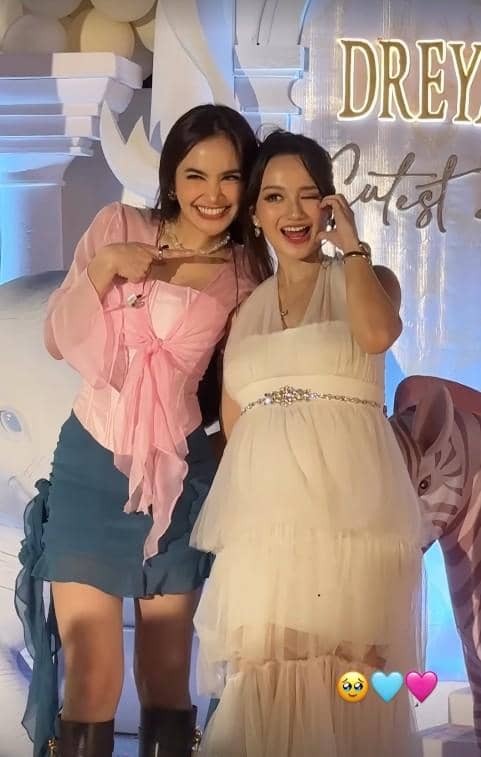 Dua wanita berpose bersama di acara gender reveal dengan latar dekorasi berwarna putih dan biru, salah satunya mengenakan gaun putih.