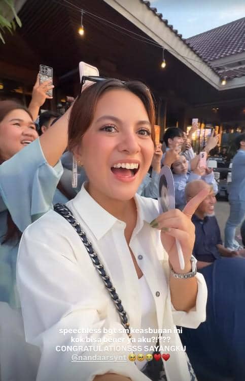 Seorang wanita tersenyum lebar sambil menunjuk ke arah depan di acara gender reveal Nanda Arsyinta, dikelilingi tamu yang memotret momen tersebut.