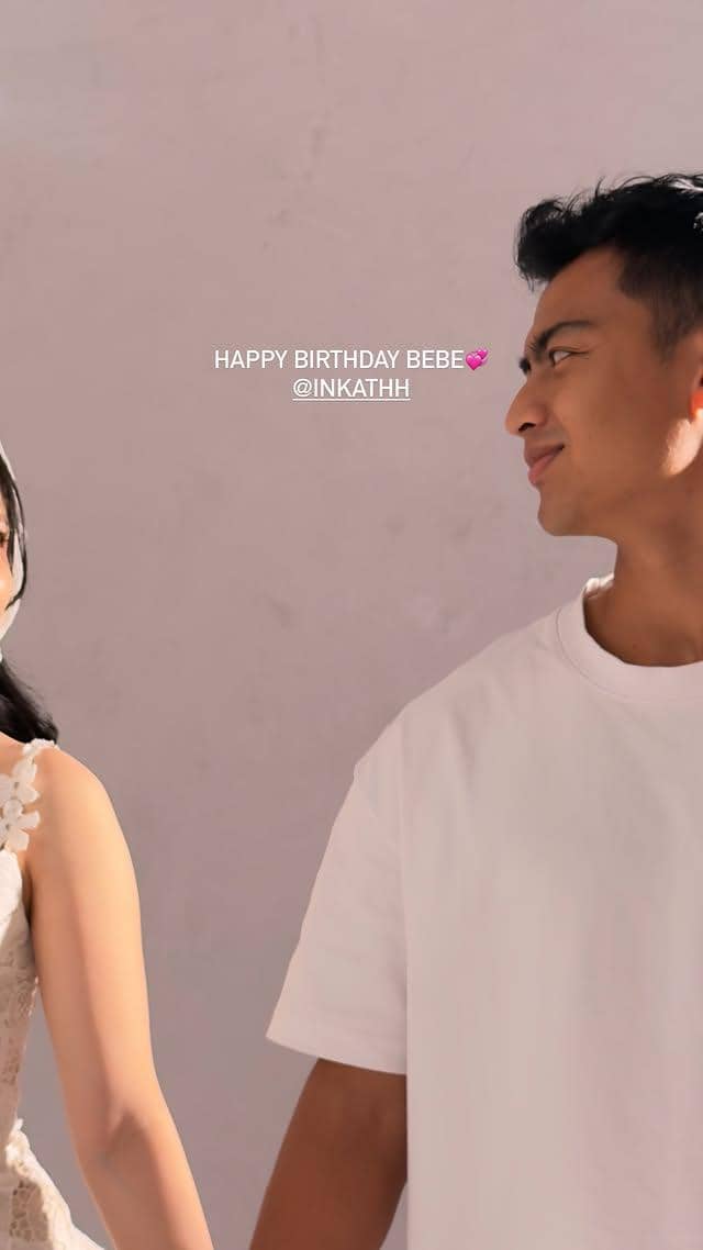 Seorang pria mengenakan kaus putih tersenyum sambil menatap wanita di sebelahnya yang mengenakan gaun putih dengan latar dinding berwarna lembut.