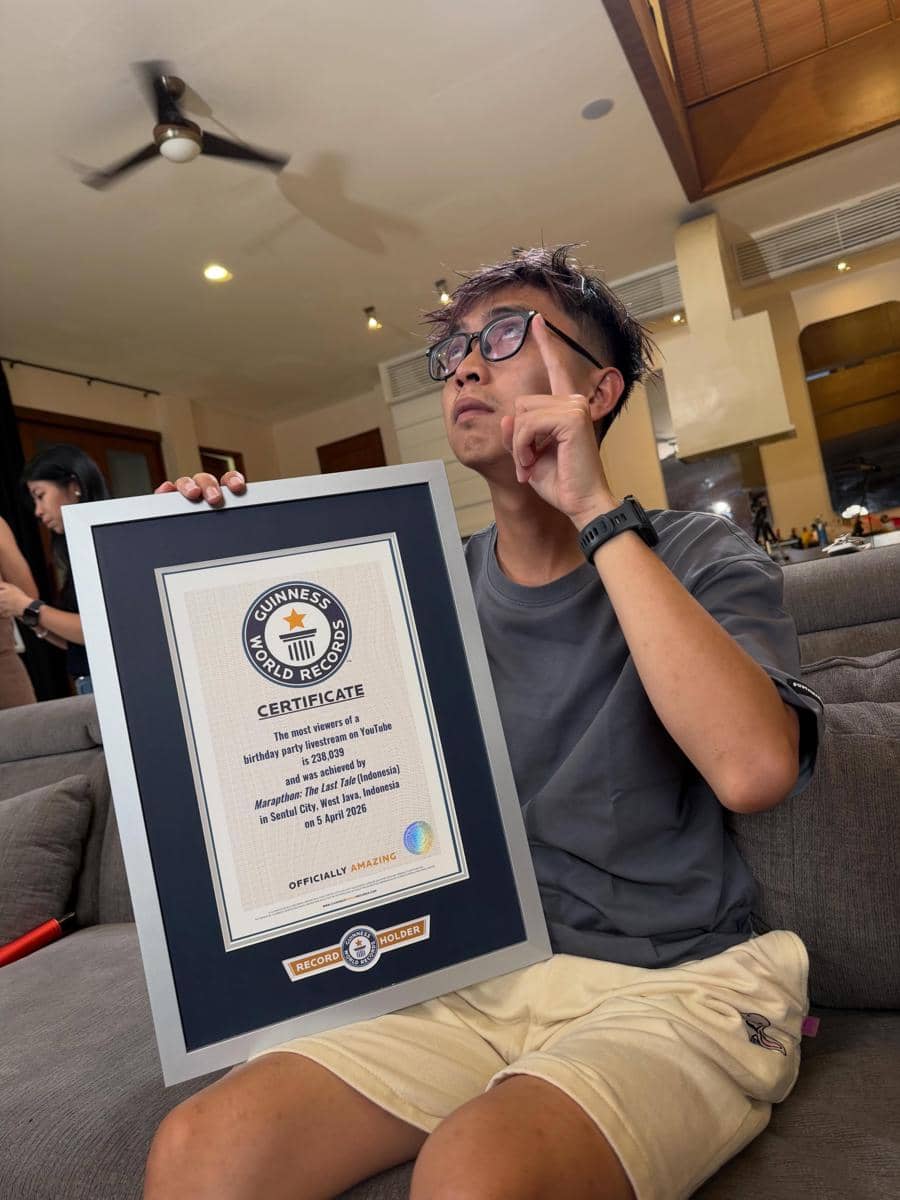 Seorang pria duduk di sofa sambil memegang sertifikat Guinness World Records berbingkai besar di ruang tamu modern.