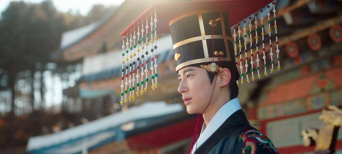 Seorang pria mengenakan hanbok kerajaan dan mahkota tradisional Korea dengan hiasan manik warna-warni di depan bangunan istana.