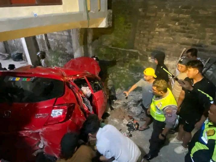 Mobil merah rusak parah setelah kecelakaan di Kupang, dikelilingi petugas kepolisian dan warga yang menolong di lokasi kejadian.
