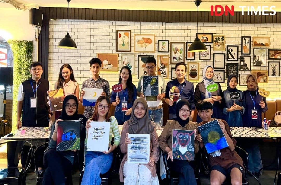 Sekelompok orang berpose bersama hasil lukisan mereka di sebuah kafe dengan dinding penuh bingkai seni dan pencahayaan hangat.