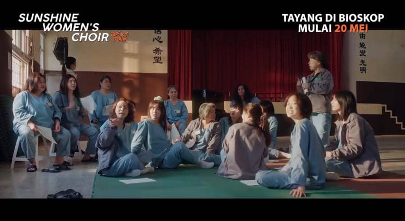 Sekelompok wanita duduk di lantai ruang latihan dengan pakaian seragam biru dalam adegan film Sunshine Women’s Choir.