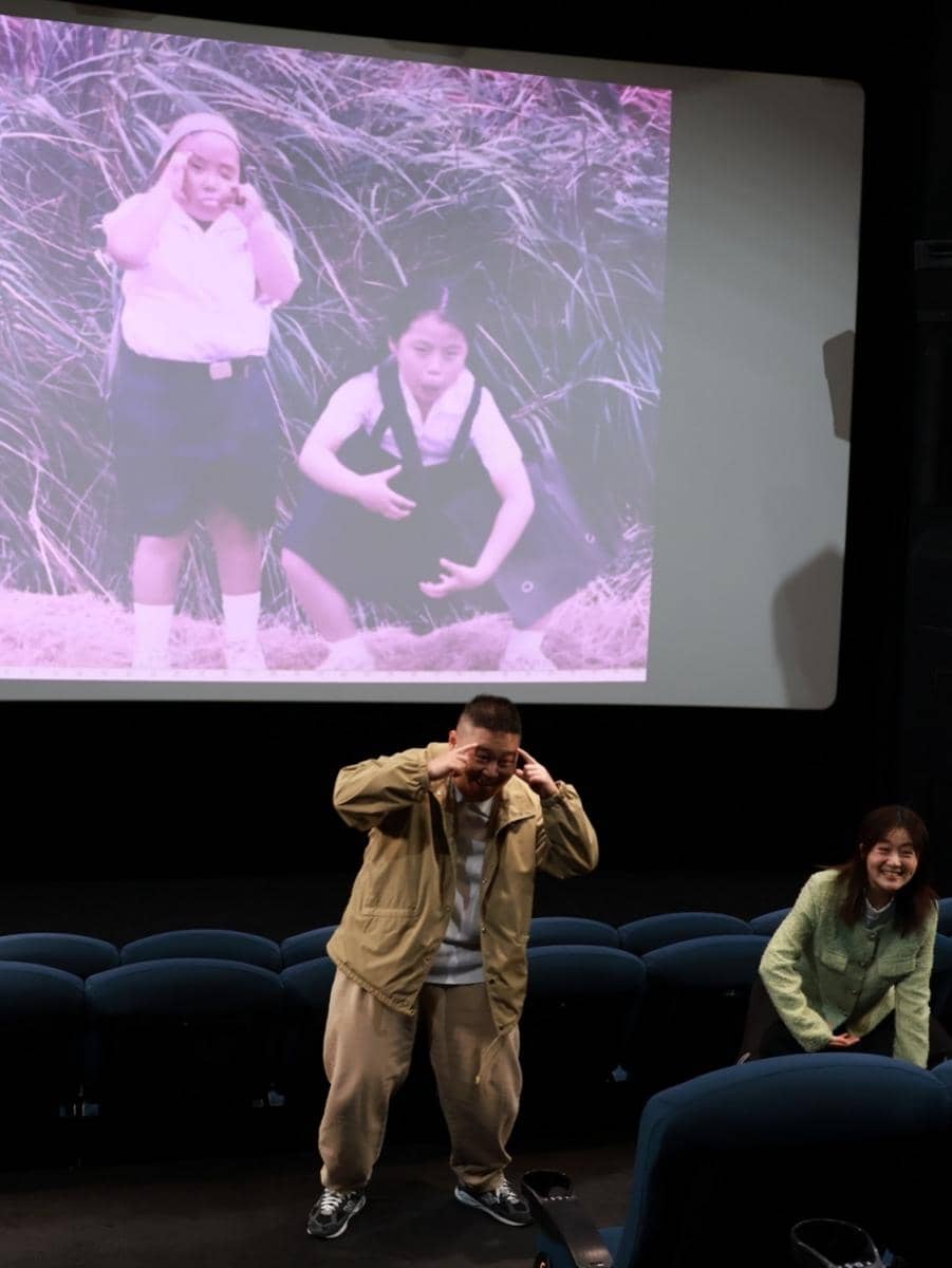 Dua orang berdiri di depan layar bioskop menirukan pose masa kecil mereka yang ditampilkan di layar di belakang.