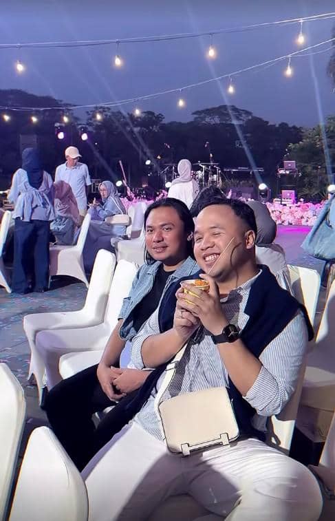 Dua pria duduk di kursi acara outdoor malam hari dengan lampu gantung dan dekorasi ungu di acara gender reveal.