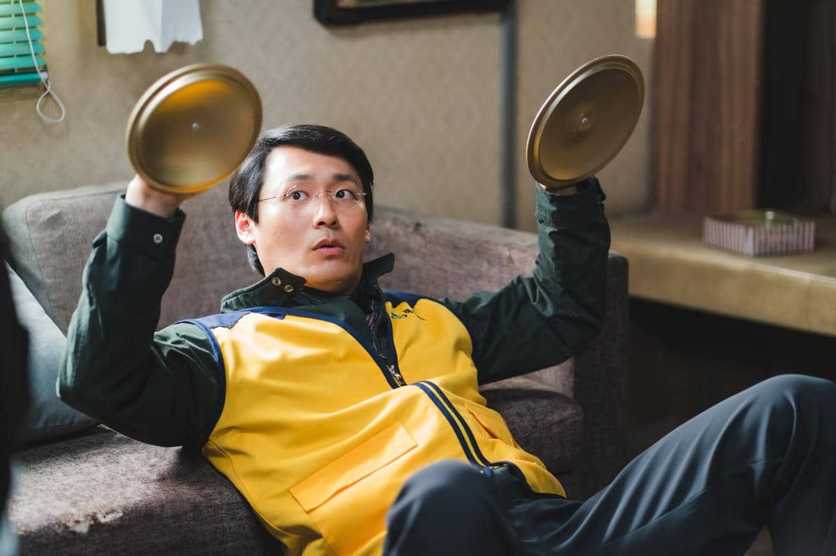 Seorang pria mengenakan rompi kuning duduk di sofa sambil memegang dua tutup panci logam dengan ekspresi terkejut di wajahnya.
