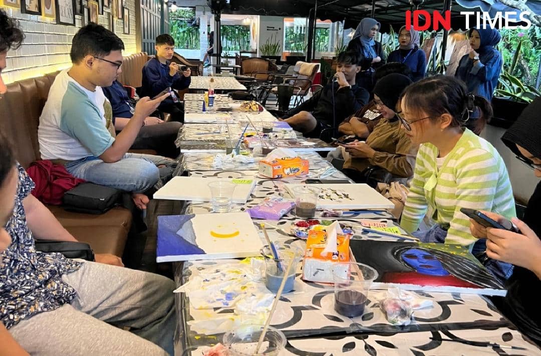 Peserta art therapy duduk di sekitar meja panjang dengan kanvas dan cat, melukis simbol serta warna yang mewakili perasaan mereka.