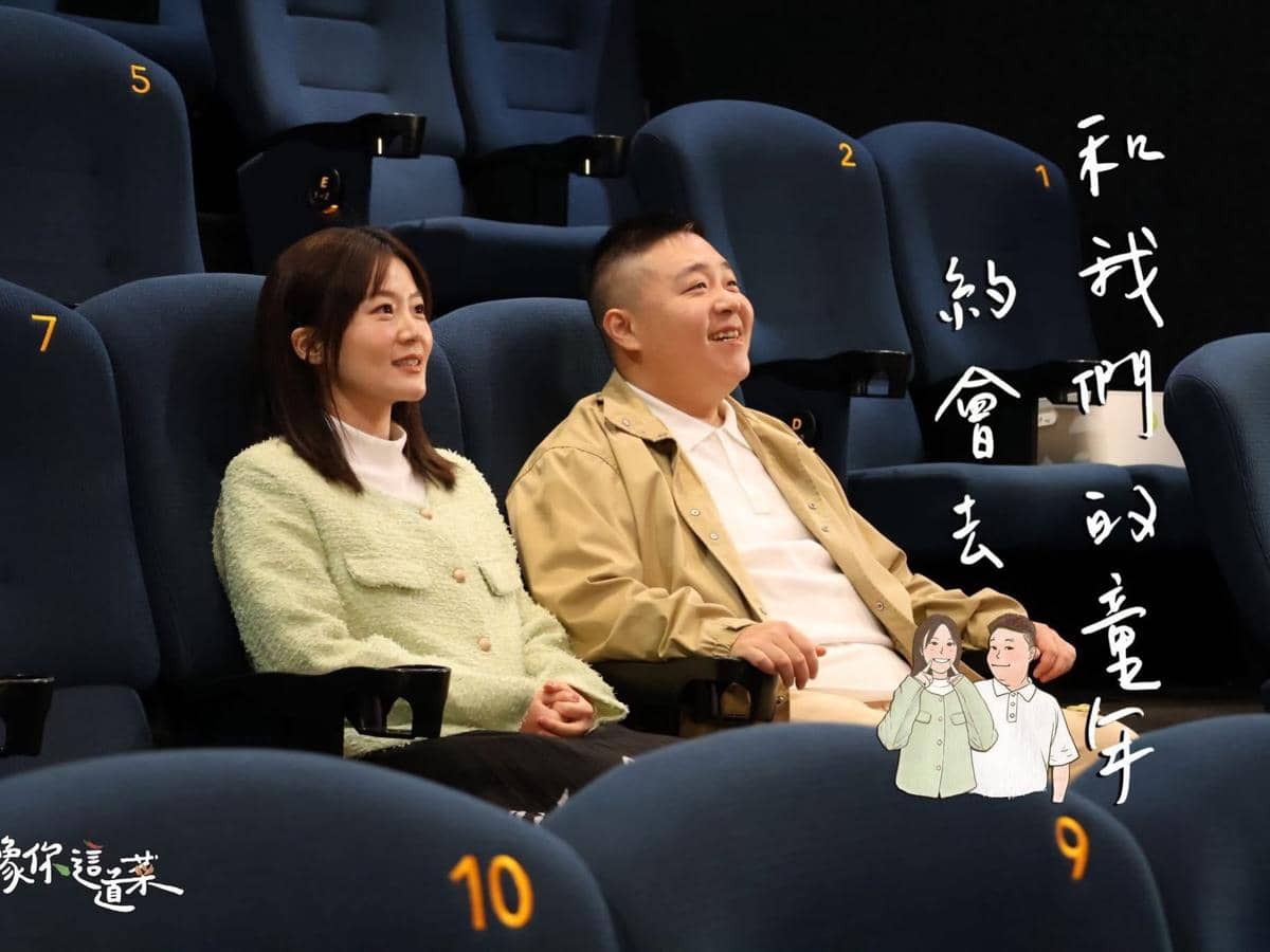 Seorang pria dan wanita duduk berdampingan di kursi bioskop bernomor, tersenyum sambil menatap ke arah layar di depan mereka.