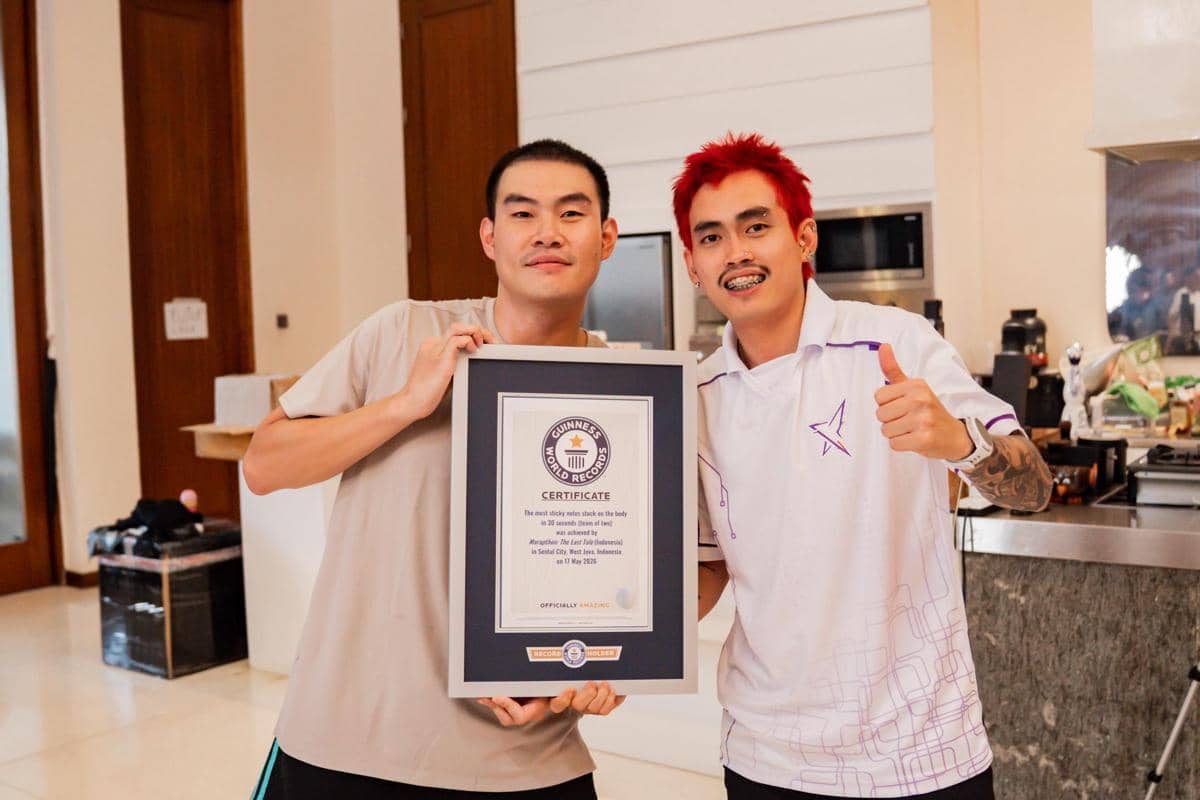 Dua pria berpose sambil memegang sertifikat Guinness World Records di dalam ruangan dengan ekspresi bahagia dan santai.