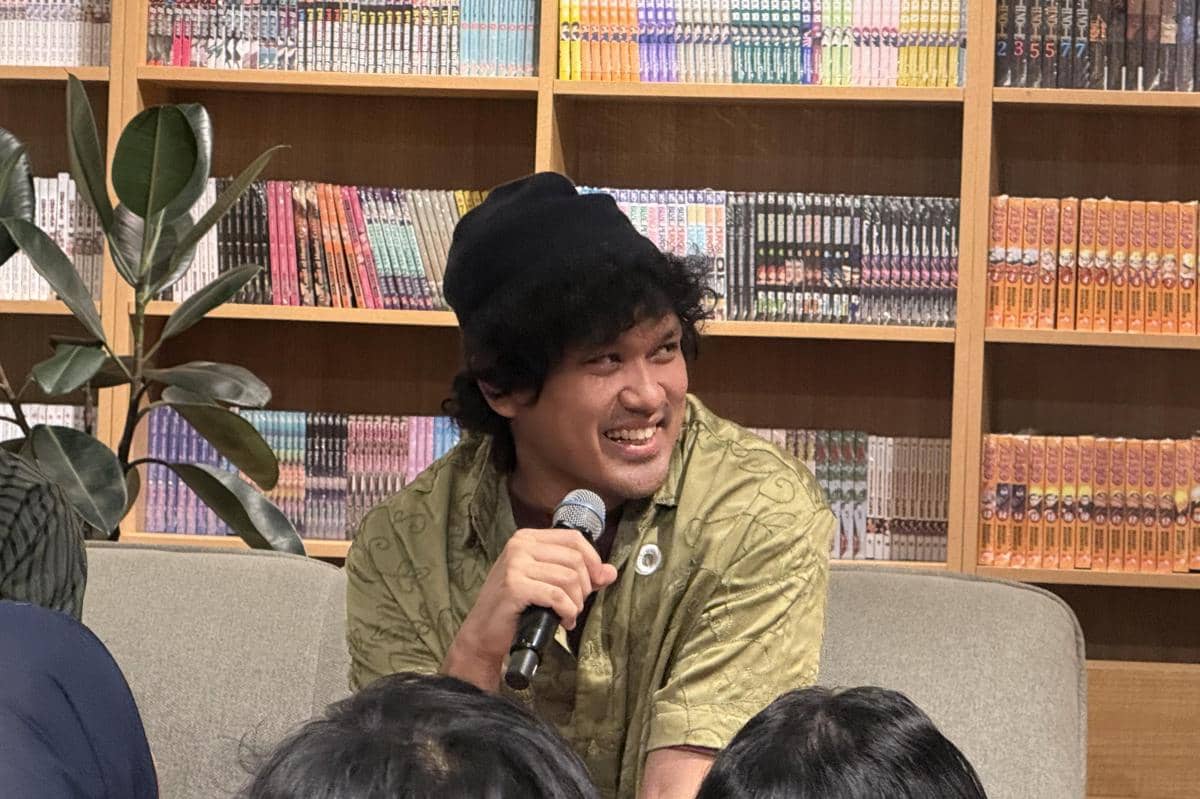 Seorang pria mengenakan kemeja hijau dan topi hitam tersenyum sambil memegang mikrofon di depan rak buku saat konferensi pers.