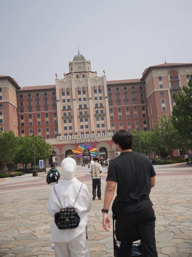 Beberapa orang berjalan menuju gerbang utama Universal Studios Beijing dengan bangunan bergaya klasik di latar belakang.