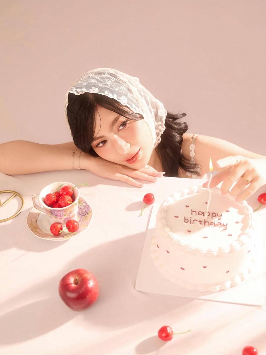 Seorang wanita mengenakan kerudung renda putih bersandar di meja dengan kue ulang tahun bertuliskan 'happy birthday' dan buah ceri di sekitarnya.